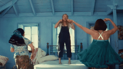 Three Women Are Jumping On A Bed With One Wearing A Green Dress GIF