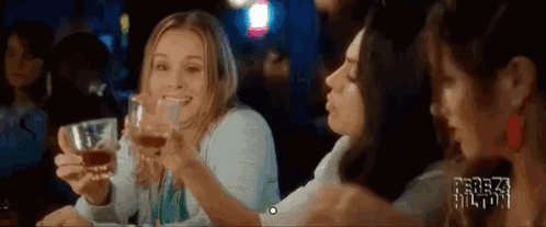 Three Women Are Sitting At A Table Holding Glasses Of Whiskey And Smiling . GIF