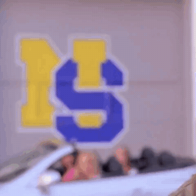 Three Women Are Sitting In A White Convertible Car In Front Of A Wall With The Letter S Painted On It . GIF