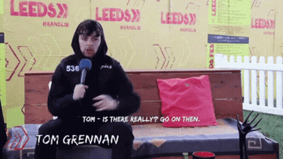 Tom Grennan Sitting On Bench With Microphone In Front Of Leeds Sign GIF