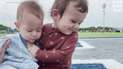 Two Babies Are Hugging Each Other On A Field And One Of Them Is Wearing A Striped Shirt GIF