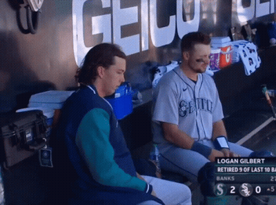 Two Baseball Players Sitting In A Dugout With Logan Gilbert On The Screen GIF