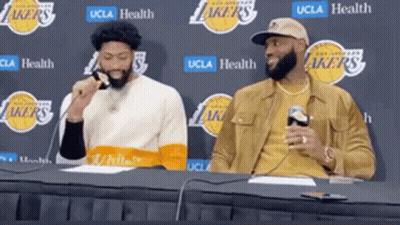 Two Basketball Players Are Sitting At A Table With A Lakers Logo Behind Them GIF