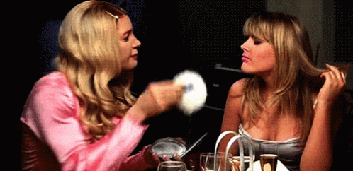 Two Blonde Women Are Sitting At A Table And One Is Holding A Powder Puff GIF