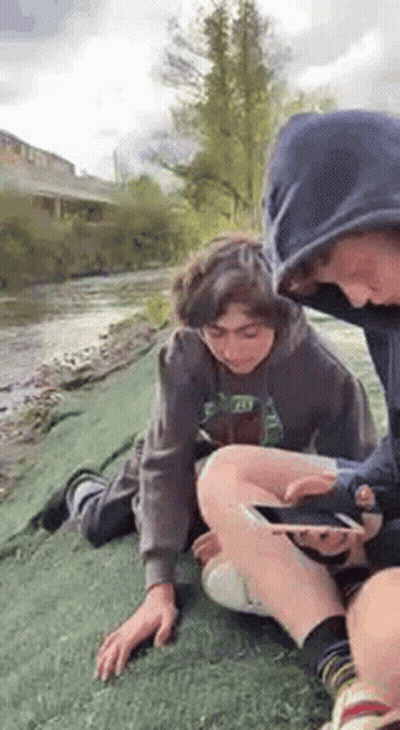 Two Boys Sitting By River Looking At Cell Phone GIF