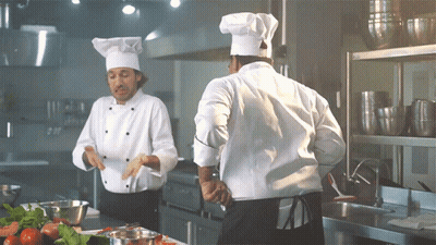 Two Chefs In Kitchen, One In White Hat GIF