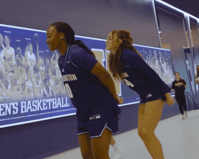 Two Female Basketball Players In Front Of Men's Basketball Wall GIF
