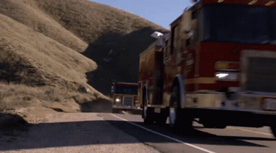 Two Fire Trucks Are Driving Down A Road With A Mountain In The Background GIF