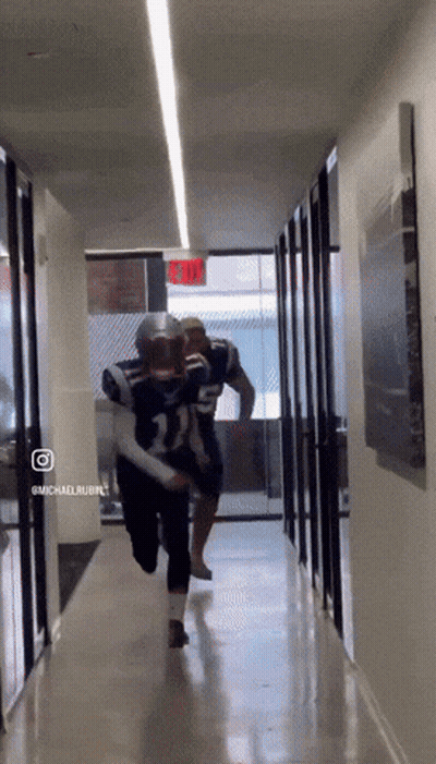 Two Football Players Are Running Down A Hallway With A Red Exit Sign Behind Them GIF