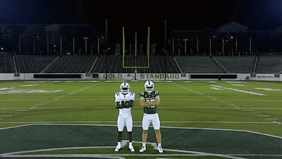 Two Football Players Standing On A Field With A Sign That Says Gold Standard In The Background GIF