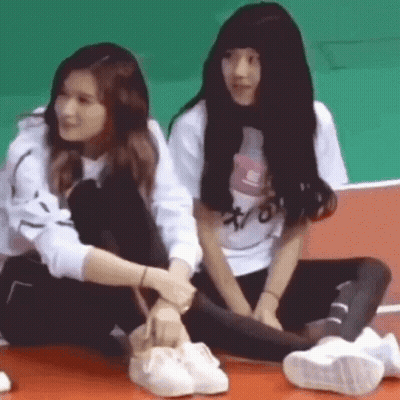 Two Girls Are Sitting Next To Each Other On The Floor And One Of Them Is Tying Another Girl 'S Shoe . GIF