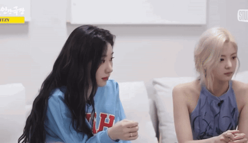 Two Girls Are Sitting On A Couch Talking To Each Other . One Of The Girls Is Wearing A Blue Sweater . GIF