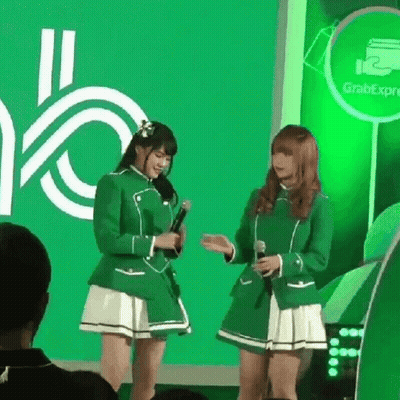 Two Girls In Green Uniforms Are Standing Next To Each Other With Microphones In Front Of A Green Background . GIF
