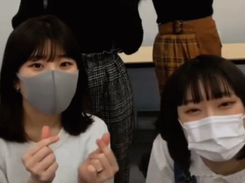 Two Girls Wearing Face Masks Are Making A Heart Shape With Their Fingers GIF