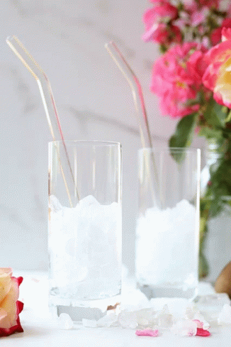 Two Glasses Filled With Ice And Straws Next To A Vase Of Flowers GIF