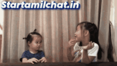 Two Little Girls Are Sitting At A Table In Front Of A Sign That Says " Starttamilchat.in " GIF
