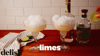 Two Margarita Glasses Filled With Ice And Limes Next To A Bottle Of Liquor GIF