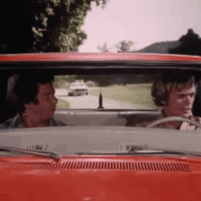 Two Men Are Driving A Red Car Down A Road . GIF