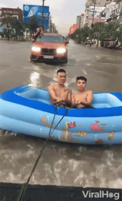 Two Men Are In An Inflatable Pool In A Flooded Street With A Car Behind Them GIF
