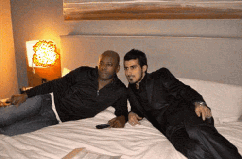 Two Men Are Laying On A Bed And One Has A Remote Control In His Hand GIF