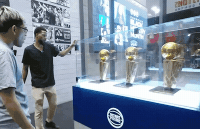 Two Men Are Looking At Trophies In A Glass Case That Says Detroit Pistons GIF