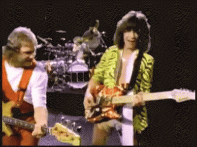 Two Men Are Playing Guitars On A Stage With A Drum Set In The Background GIF