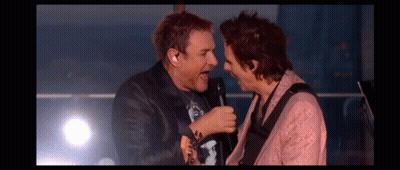 Two Men Are Singing Into Microphones On A Stage While Playing Guitars . GIF