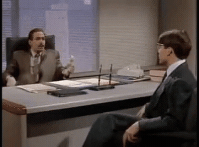 Two Men Are Sitting At A Desk Talking To Each Other . GIF