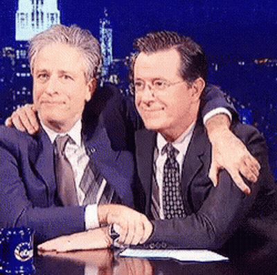 Two Men Are Sitting At A Table And One Has His Arm Around The Other 'S Shoulders GIF