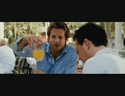 Two Men Are Sitting At A Table And One Is Pouring Orange Juice Into Another Man 'S Glass GIF