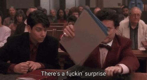 Two Men Are Sitting At A Table And One Of Them Is Holding A Piece Of Paper That Says There 'S A Fuckin Surprise GIF