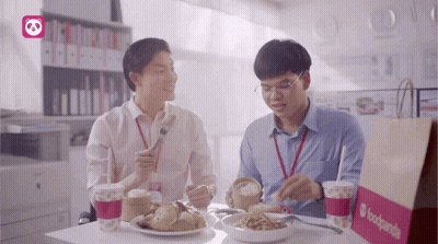 Two Men Are Sitting At A Table Eating Food And Drinking From A Bag That Says Foodpanda GIF