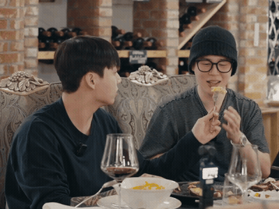Two Men Are Sitting At A Table Eating Food And Drinking Wine With Bottles Of Wine In The Background GIF