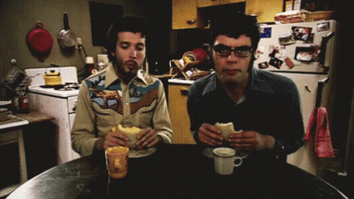 Two Men Are Sitting At A Table Eating Sandwiches In Front Of A Refrigerator With Pictures On It GIF