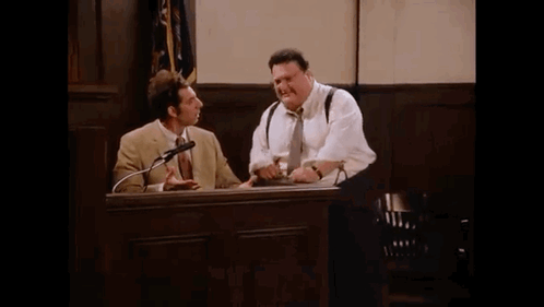 Two Men Are Sitting At A Table In A Courtroom Talking Into Microphones . GIF