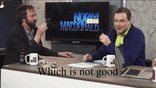 Two Men Are Sitting At A Table In Front Of A Norm Macdonald Tv GIF