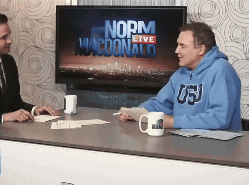 Two Men Are Sitting At A Table In Front Of A Screen That Says Norma Macdonald GIF