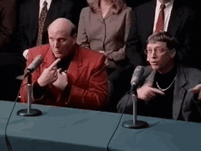 Two Men Are Sitting At A Table Talking Into Microphones While A Group Of People Watch . GIF