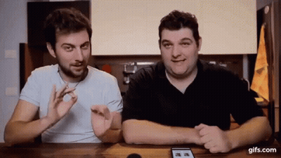 Two Men Are Sitting At A Table Talking To Each Other And Looking At A Phone . GIF