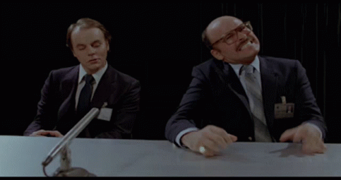Two Men Are Sitting At A Table With A Microphone And One Has A Name Tag That Says ' A. ' GIF