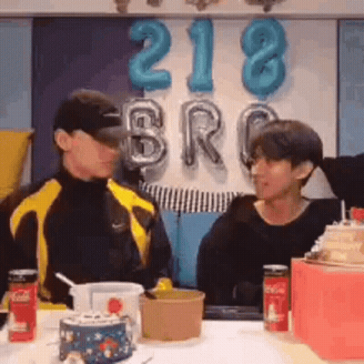 Two Men Are Sitting At A Table With Balloons In The Background And Talking To Each Other . GIF