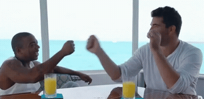 Two Men Are Sitting At A Table With Glasses Of Orange Juice And Fist Bumps GIF