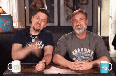 Two Men Are Sitting At A Table With One Wearing A Shirt That Says Dragons On It GIF