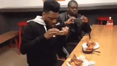 Two Men Are Sitting At A Table With Plates Of Food And One Is Taking A Picture Of The Other . GIF