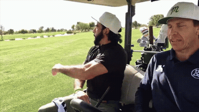 Two Men Are Sitting In A Golf Cart With One Wearing A White Hat With The Letter M On It GIF