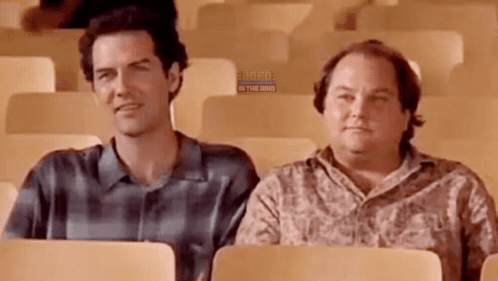 Two Men Are Sitting In A Row Of Chairs In A Stadium . GIF