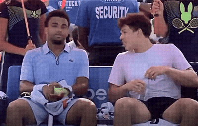 Two Men Are Sitting In A Stadium With A Security Guard In The Background GIF