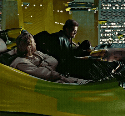 Two Men Are Sitting In A Yellow And Green Vehicle With A City In The Background GIF