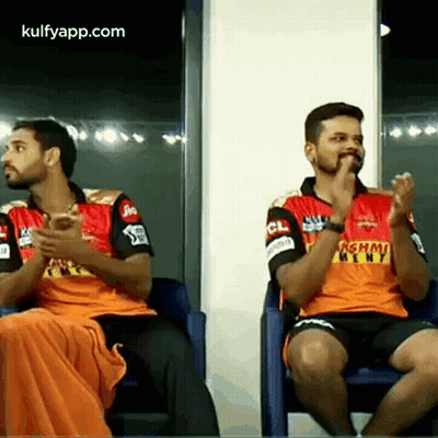 Two Men Are Sitting In Chairs And Clapping Their Hands While Wearing Orange Jerseys GIF