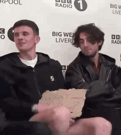 Two Men Are Sitting In Front Of A Big Web Live Sign GIF